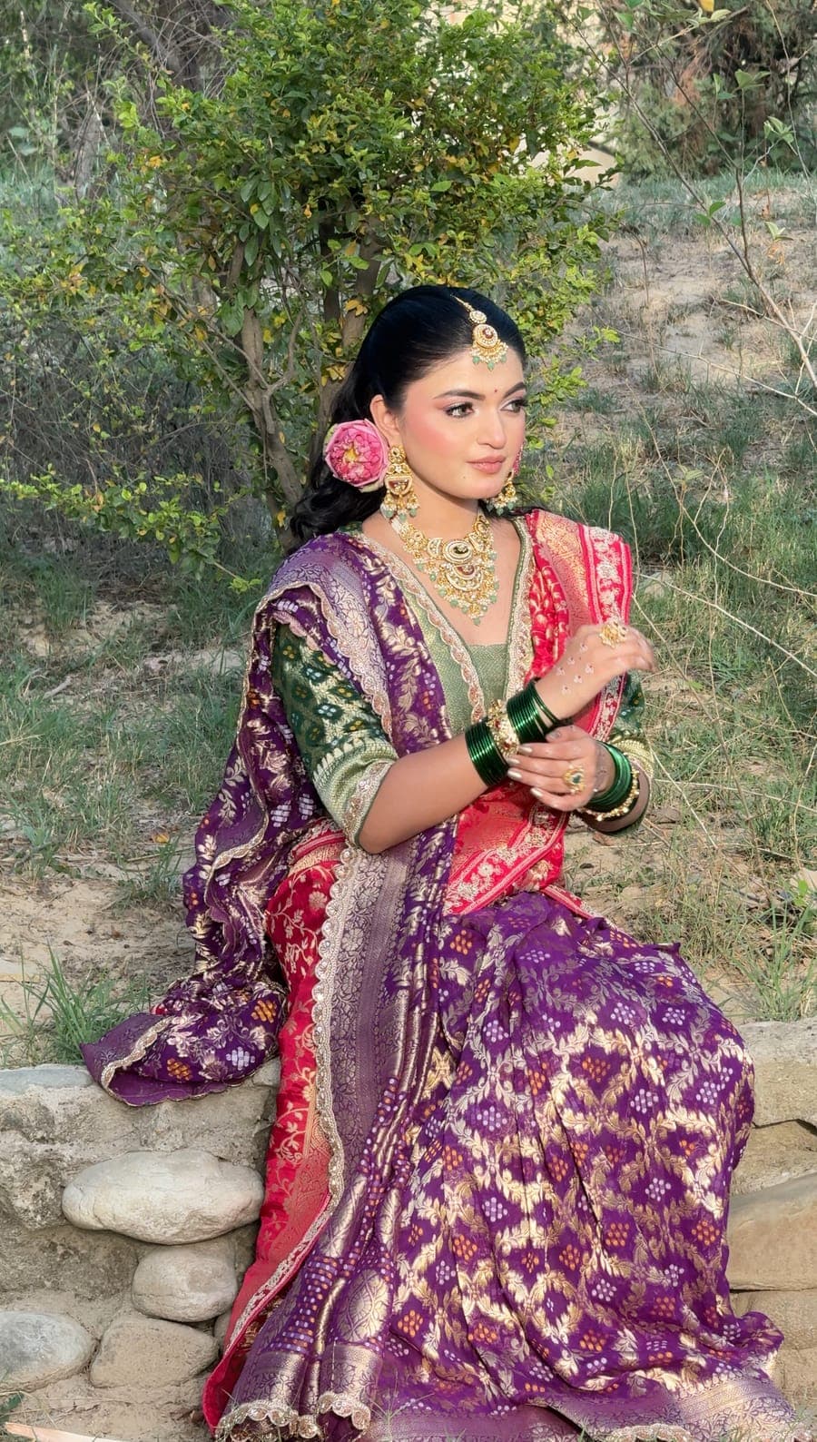 Bride in a purple lehenga seated beside a stone wall with green bangles